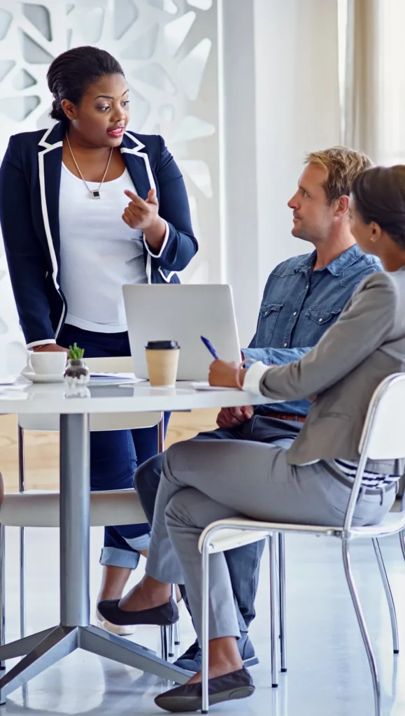 A female professional stands and speaks to two people seated at a table